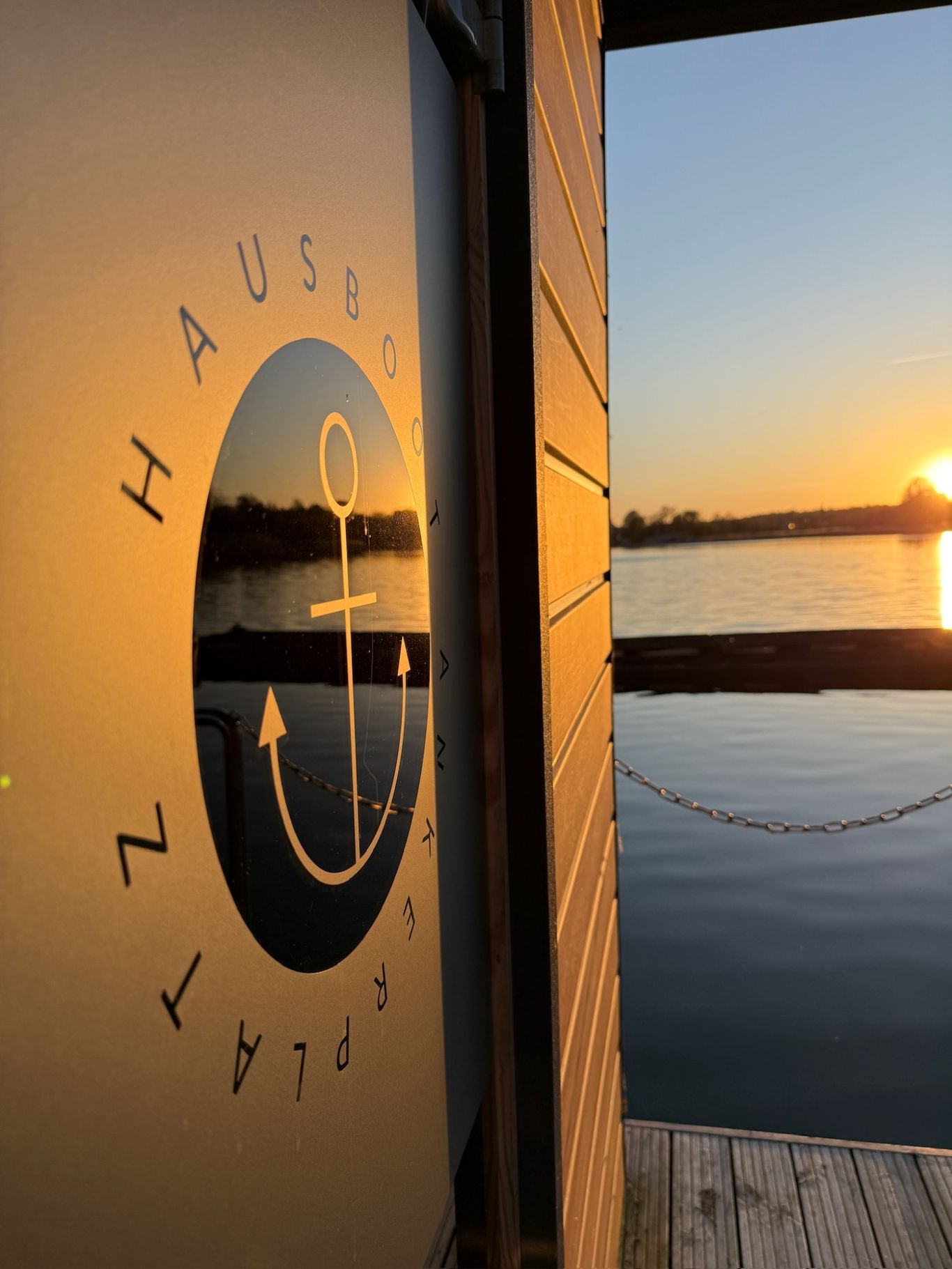 Im Linken Bildrand sieht man die Saunatür mit dem Logo von Hausboot Ankerplatz. Rechts im Bild die untergehende Sonne. Wasserspiegelung.